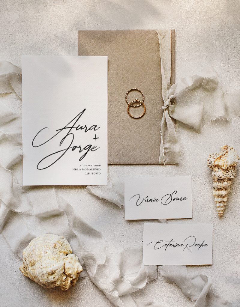 Convite de casamento de estilo tipográfico com um laço de linho bege, com um envelope cinza pousado numa bandeja dourada