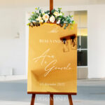 Placa de boas vindas em acrílico dourado num cavalete com detalhes florais